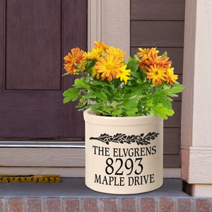 May include: A cream-colored ceramic planter with black lettering that reads "THE ELVGRENS 8293 MAPLE DRIVE". The planter is filled with a green plant with orange flowers.