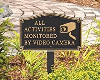 May include: A black metal sign with gold lettering that reads "ALL ACTIVITIES MONITORED BY VIDEO CAMERA". A small black and white camera icon is on the sign. The sign is mounted on a black metal post and is in a garden setting.