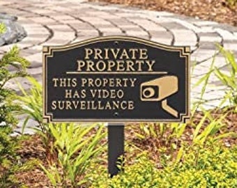 Peut inclure: Un panneau en métal noir avec des lettres dorées qui indiquent "Propriété privée Cette propriété est sous vidéo surveillance". Le panneau comporte une petite image d'une caméra de sécurité dans le coin supérieur droit. Le panneau est monté sur un poteau en métal noir et est placé dans un jardin.