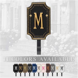 May include: Black and gold metal address sign with the letter "M". The sign has a decorative border and is mounted on a black stake. The text "11 COLORS AVAILABLE" is displayed below the sign, with examples of the available colors.