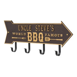 May include: A brown metal sign with gold lettering and a directional arrow shape. The sign reads "Uncle Steve's World Famous BBQ" and includes a fork and spatula graphic. It has four black hooks below.