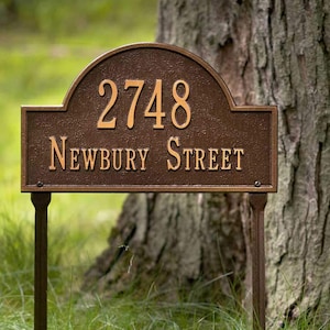 May include: A bronze-colored house number sign with the number 2748 and the text "NEWBURY STREET" in gold lettering. The sign is mounted on two metal stakes and is in front of a tree.