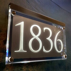 May include: A white acrylic house number sign with the number 1836 on a brown background. The sign is framed with a silver metal frame and has silver screws.