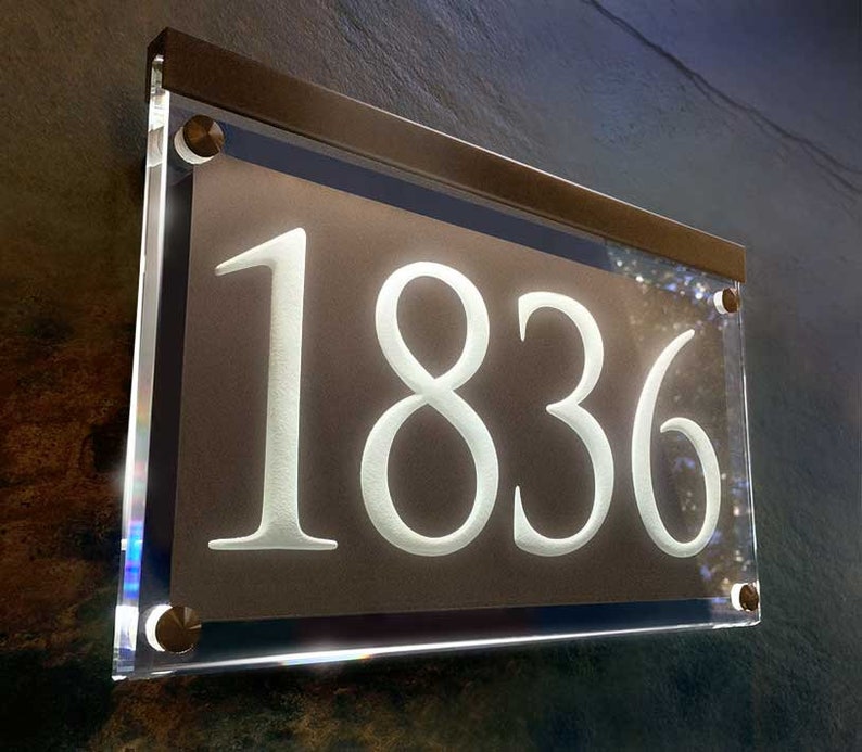 May include: A white acrylic house number sign with the number 1836 on a brown background. The sign is framed with a silver metal frame and has silver screws.
