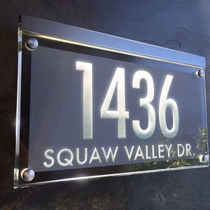 May include: A modern house number sign with a silver frame and clear glass. The number 1436 is displayed in white on a dark gray background. The street name "SQUAW VALLEY DR." is also displayed in white.