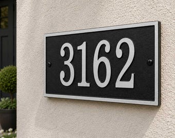 Puede incluir: Una placa rectangular de número de casa con los dígitos 3162 en relieve de color plateado sobre un fondo negro texturizado. La placa tiene un marco plateado y está fijada a la pared con dos tornillos negros visibles a la izquierda y derecha.