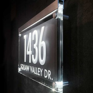 May include: A clear acrylic house number sign with a brushed silver frame. The sign displays the number "1436" in white lettering and the street address "SQUAW VALLEY DR." in white lettering.