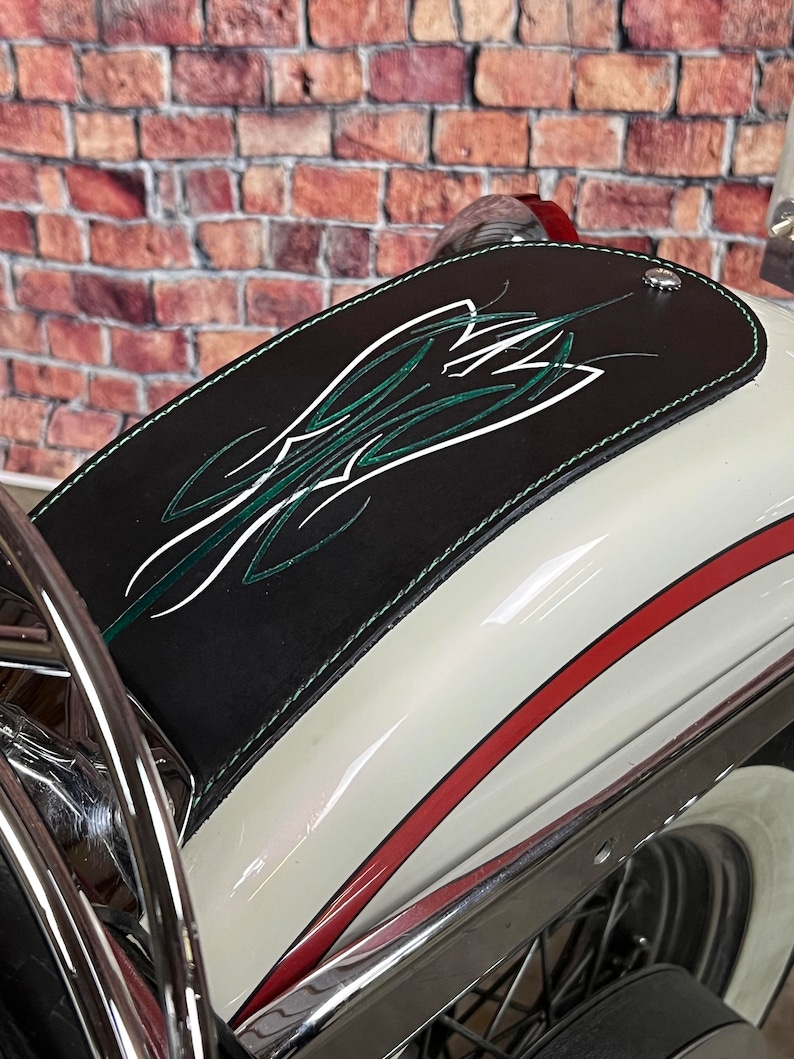 K&ouml;nnte beinhalten: Ein wei&szlig;er Motorradkotfl&uuml;gel mit einer schwarzen Lederabdeckung. Die Abdeckung hat ein gr&uuml;n-wei&szlig;es Pinstripe-Design. Der Kotfl&uuml;gel hat einen roten Streifen am Rand.