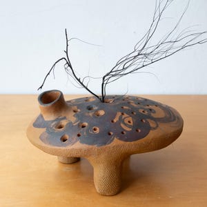 May include: A handcrafted, terracotta-colored ceramic vase with a unique, mushroom-like design. The vase features a flat, perforated top with dark brown accents and three sturdy legs. A sprig of dark, dried branches rises from the center, adding a natural element.