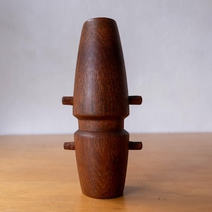 Könnte beinhalten: Eine handgefertigte, dunkelbraune Holzvase mit einem einzigartigen, geschichteten Design. Die Vase hat eine konische Oberseite und einen breiteren Sockel mit kleinen, horizontalen Holzvorsprüngen an den Seiten. Die Holzmaserung ist sichtbar.