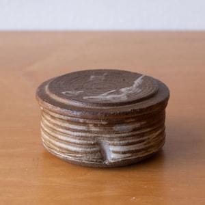 May include: A round, lidded ceramic container with a dark brown, textured finish. The exterior features horizontal ridges along the sides and concentric circular grooves on the lid, with subtle white glaze accents highlighting the clay texture.