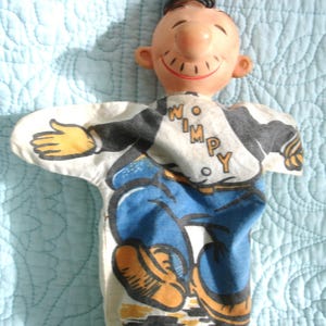 May include: A vintage hand puppet featuring a smiling cartoon character with a brown hat and a black and white shirt with the word "WIMPY". The puppet's pants are blue, and shoes are orange. The puppet is made of fabric and plastic.