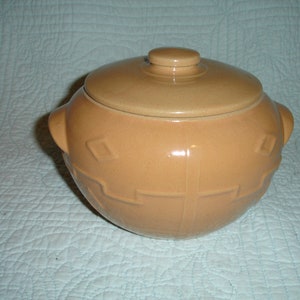 May include: A tan ceramic casserole dish with a lid. The dish has two handles and a decorative geometric pattern around the base. The lid has a small knob handle. The dish is likely used for cooking and serving food.