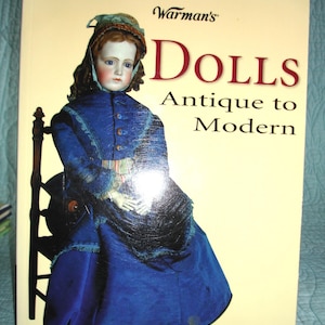 May include: A book titled "Warman's Dolls Antique to Modern" features a doll in a blue dress and bonnet. The book cover has a cream background with red and black text. The book is an identification and price guide.
