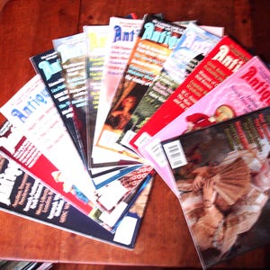 May include: A collection of Antique Doll Collector magazines arranged in a fan shape. Each magazine cover features the title Antique Doll Collector in bold lettering, along with various photographs of antique dolls and descriptive text headlines.