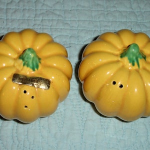May include: A pair of ceramic salt and pepper shakers shaped like pumpkins. The shakers are a bright yellow with green stems and have small holes for dispensing. The label on one shaker reads "HEADLANDS".