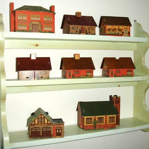 Puede incluir: Una colecci&oacute;n de casas de papel en miniatura de varios estilos, exhibidas en una estanter&iacute;a de madera verde claro de tres niveles. Las casas presentan dise&ntilde;os de ladrillo, piedra y tejas, con detalles como chimeneas y ventanas.