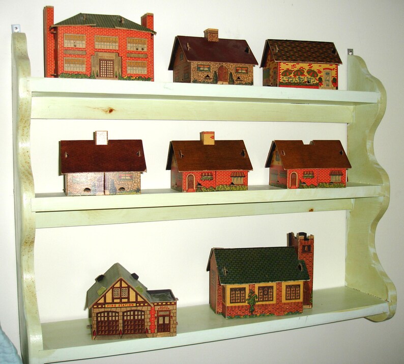 Puede incluir: Una colecci&oacute;n de casas de papel en miniatura de varios estilos, exhibidas en una estanter&iacute;a de madera verde claro de tres niveles. Las casas presentan dise&ntilde;os de ladrillo, piedra y tejas, con detalles como chimeneas y ventanas.