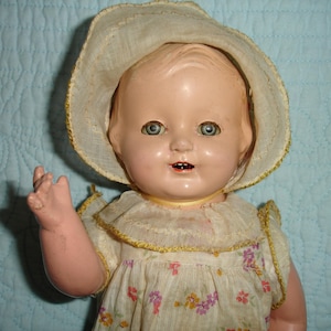 May include: Vintage baby doll with blue eyes, wearing a floral print dress and a matching sun hat. The doll has a painted face and open mouth, with a yellow ribbon around its neck. The doll's arm is raised in a waving gesture.