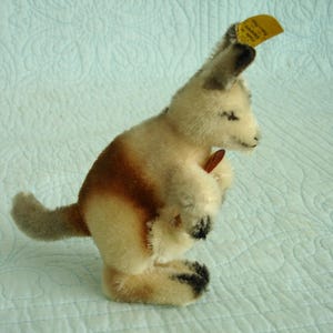 May include: A vintage plush kangaroo toy in cream, brown, and black. The kangaroo has a small pouch and a tag attached to its ear. The toy is made of soft, plush material and is posed standing upright.