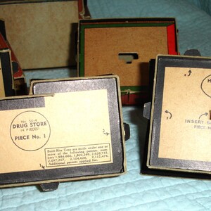 Puede incluir: Se muestran varias cajas de juguetes antiguos, de forma rectangular con bordes negros y etiquetas de papel color beige. Las etiquetas incluyen texto como "Drug Store" y "House No. 5", indicando el contenido. Las cajas est&aacute;n en varios estados de montaje.