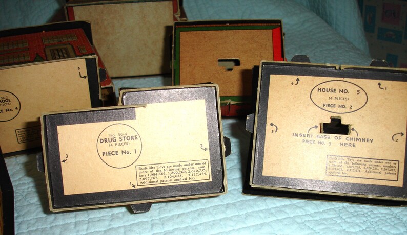 Puede incluir: Se muestran varias cajas de juguetes antiguos, de forma rectangular con bordes negros y etiquetas de papel color beige. Las etiquetas incluyen texto como "Drug Store" y "House No. 5", indicando el contenido. Las cajas est&aacute;n en varios estados de montaje.