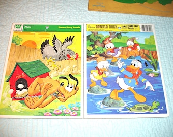 Könnte beinhalten: Zwei Vintage-Rahmenpuzzles mit Disney-Figuren. Das linke Puzzle zeigt den Hund Pluto bei einer roten Hundehütte und einem Huhn. Das rechte Puzzle zeigt Donald Duck und drei Entenküken an einem Teich mit einer grünen Schildkröte.