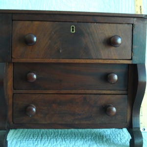 May include: A dark brown wooden chest of drawers with three horizontal drawers, each featuring two round knobs. The top drawer includes a central brass keyhole escutcheon. A yellow ruler is visible on the right side, showing measurements in inches.
