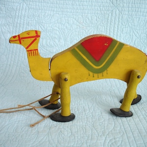 May include: A vintage wooden pull-along camel toy. The camel is painted yellow with red and green accents on the saddle and head. It has four legs with black feet and a string for pulling. A classic toy.