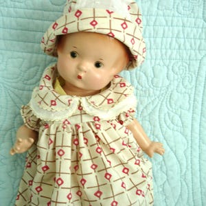 May include: Vintage doll wearing a patterned dress and matching hat. The dress and hat feature a cream background with a repeating red diamond and brown cross design. The doll has a painted face and is posed on a light blue quilted surface.