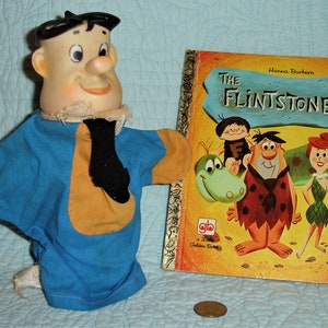 May include: A vintage Fred Flintstone hand puppet with a blue and yellow shirt, black tie, and a smiling face. Beside it is a vintage copy of "The Flintstones" children's book with cartoon characters on the cover.