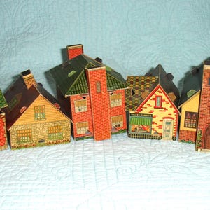 Puede incluir: Una colecci&oacute;n de casas de papel en miniatura y coloridas. Las casas presentan tejados de ladrillo, verdes y marrones, con detalles como ventanas, puertas y chimeneas. Las casas est&aacute;n dispuestas en fila, creando una encantadora escena de pueblo.
