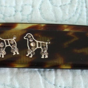May include: A tortoiseshell-patterned hair clip with two silver-colored poodle dog designs. Each dog is embellished with clear rhinestones. The clip has a rounded shape with a metal clasp.