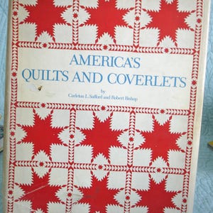 May include: A vintage book titled "America's Quilts and Coverlets" with a red and white star pattern on the cover. The book is held in a metal stand. The title is in blue font.