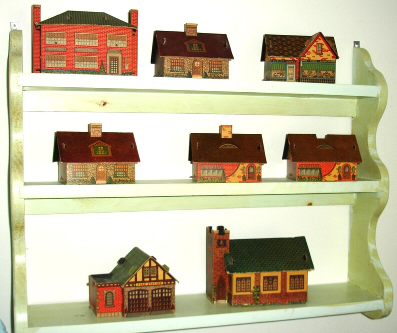 Puede incluir: Una colecci&oacute;n de casas de papel en miniatura de varios estilos, exhibidas en una estanter&iacute;a de tres niveles de color verde claro. Las casas presentan dise&ntilde;os de ladrillo, piedra y madera con ventanas, puertas y chimeneas detalladas. La estanter&iacute;a est&aacute; montada en una pared blanca.