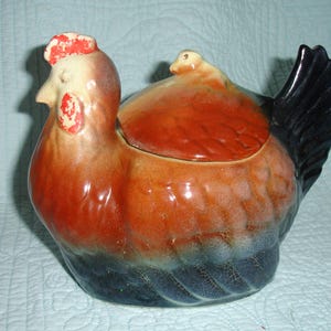May include: A ceramic chicken-shaped cookie jar. The jar is primarily orange and brown, with a black tail and a red comb. The lid is integrated into the chicken's body, and a small chick is perched on top. The jar has a glossy finish and a vintage aesthetic.