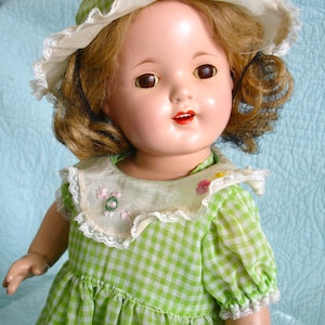 May include: A vintage doll with brown eyes, red lips, and curly brown hair. The doll wears a green and white checkered dress and a matching hat with a white lace brim. The dress has a white lace collar with embroidered flowers.