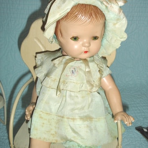 May include: Vintage doll with green eyes, red lips, and light brown hair, wearing a light green dress, bloomers, and a matching bonnet. The doll is seated in a cream-colored chair. The doll's height is approximately 24 inches.
