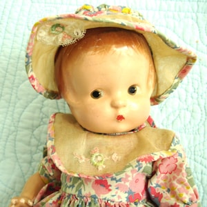 May include: A vintage doll with a painted face, wearing a floral print dress and a matching sun hat. The hat has a lace trim and is adorned with small yellow accents. The doll's dress features a pink, blue, and green floral pattern.