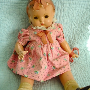 May include: Vintage doll with a painted face, wearing a pink striped dress with blue floral accents and a white collar. The doll has a matching pink bow on its head and is wearing black shoes and beige socks.