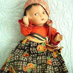 May include: A vintage doll wearing a red top and a floral patterned skirt. The doll has a red floral head covering and is posed with one arm raised. The skirt is black with yellow and red roses.