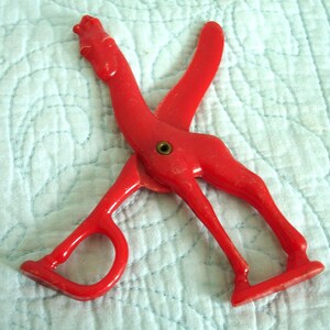 May include: A vintage red plastic giraffe-shaped scissor. The scissor has a brass rivet and is resting on a light blue quilted surface. The giraffe's head forms the top of the scissor, and the legs form the handles.