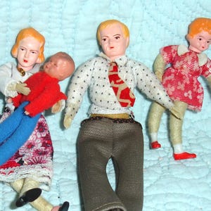 May include: A group of vintage dollhouse dolls. A woman doll in a white top and floral skirt holds a baby doll in a red jacket and blue pants. A man doll wears a polka dot shirt and brown pants. A child doll wears a red dress.