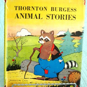 May include: Vintage children's book titled "Thornton Burgess Animal Stories." The cover features a cartoon raccoon in blue pants and a black jacket, holding a walking stick. The background is a yellow sky with trees and a grassy area.