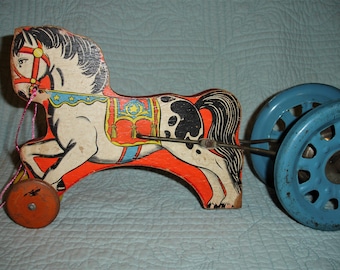 May include: Vintage wooden pull toy in the shape of a horse. The horse is painted white with black spots and a black mane. It has a red and orange body with blue wheels and a pink pull string. A classic toy.