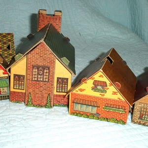 Puede incluir: Una colecci&oacute;n de casas de papel vintage con fachadas detalladas de ladrillo y piedra, tejados verdes y toldos decorativos. Una casa presenta el texto "Drugs - Sodas - Confectionery". Las casas est&aacute;n dispuestas sobre una superficie acolchada blanca.
