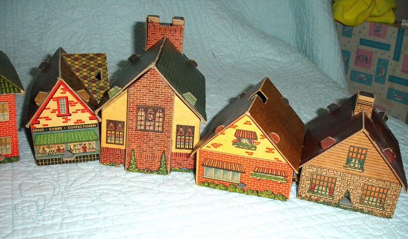 Puede incluir: Una colecci&oacute;n de casas de papel vintage con fachadas detalladas de ladrillo y piedra, tejados verdes y toldos decorativos. Una casa presenta el texto "Drugs - Sodas - Confectionery". Las casas est&aacute;n dispuestas sobre una superficie acolchada blanca.