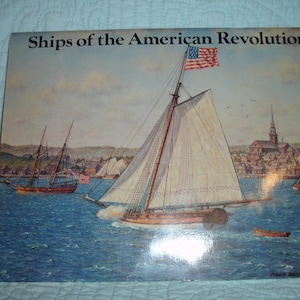 May include: A book cover titled "Ships of the American Revolution" featuring a painting of several sailing ships on the water. The ships have white sails and are set against a backdrop of a town with a church. An American flag flies from one ship.