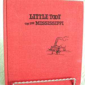 May include: A vintage red book titled "Little Toot on the Mississippi." The cover features black text and a black illustration of a steamboat. The book is displayed in a silver metal book stand.