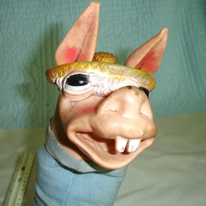 May include: A rubber donkey hand puppet featuring a tan face with large black eyes, prominent white teeth, and pink-tinted ears. The donkey wears a yellow textured hat. A light blue fabric sleeve extends from the base, next to a ruler showing inches.